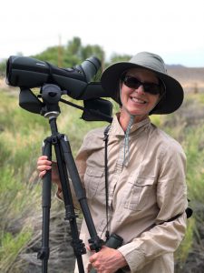 Jennifer Jones, smiling, wearing sunglasses and a large brim hat, holding a large camera on tripod